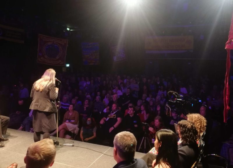 Beth Winter on a stage speaking to a crowd of around 500 people in a theatre in Liverpool. On stage with Beth are other Socialist Campaign group MPs.
